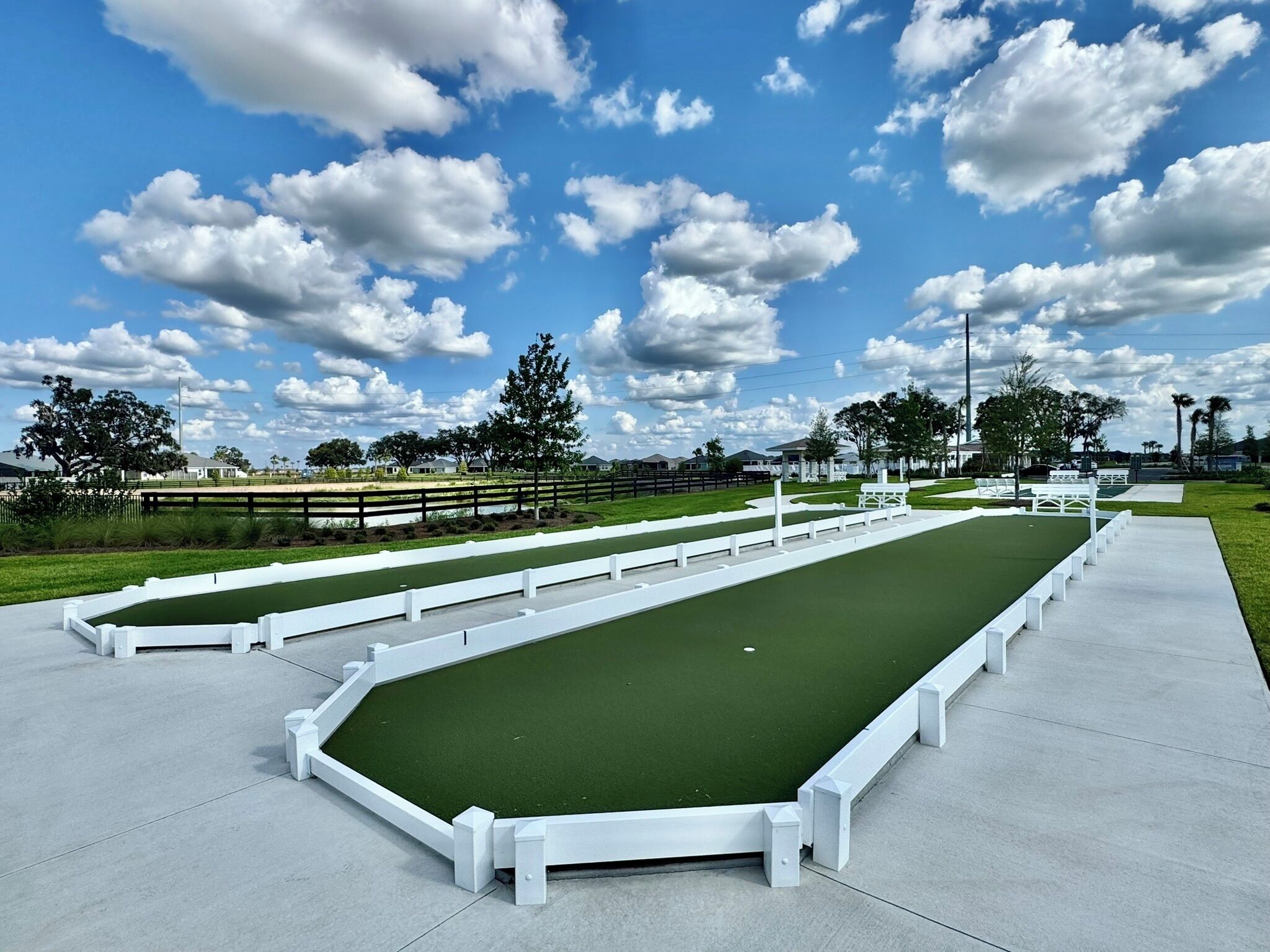 Un terrain de croquet avec des barrières blanches et une surface de jeu verte, entouré d’un chemin en béton et d’arbres.