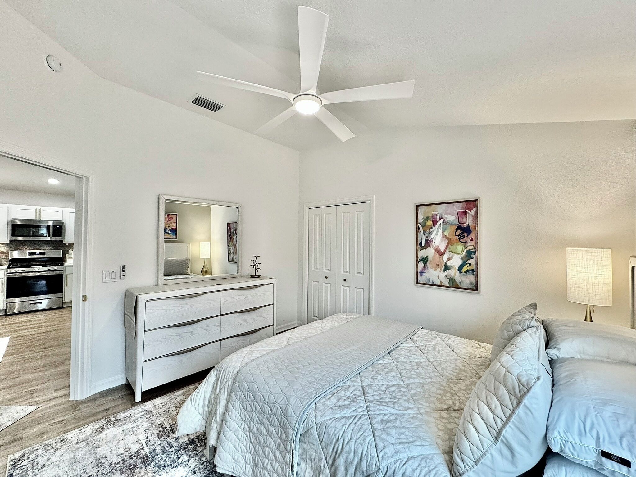 A bedroom with a bed, a dresser, a ceiling fan, a microwave, and a stove in the adjacent kitchen.
