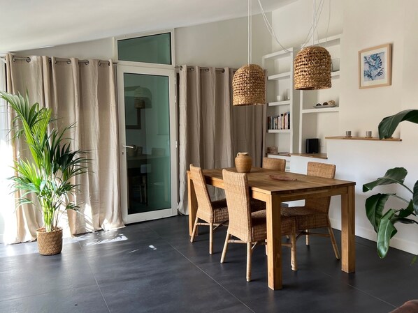 A modern dining area with a wooden table and wicker chairs, a potted plant, and a glass door leading to another room.