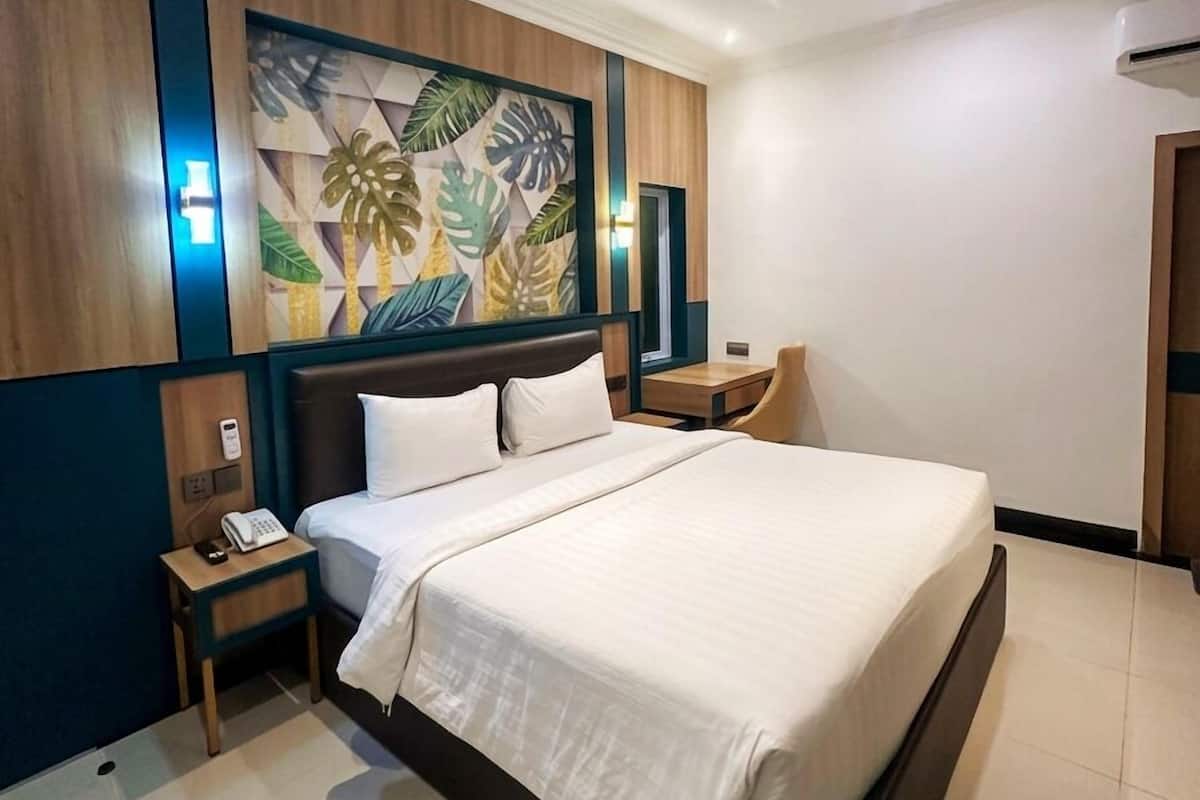 Quarto de hotel moderno com uma cama grande, uma mesa de cabeceira com telefone, uma luminária fixada na parede e uma obra de arte com tema tropical.