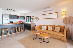 Een moderne woonkamer met een bank, twee ronde tafels, houten krukken, een keukenhoek en een grote spiegel.