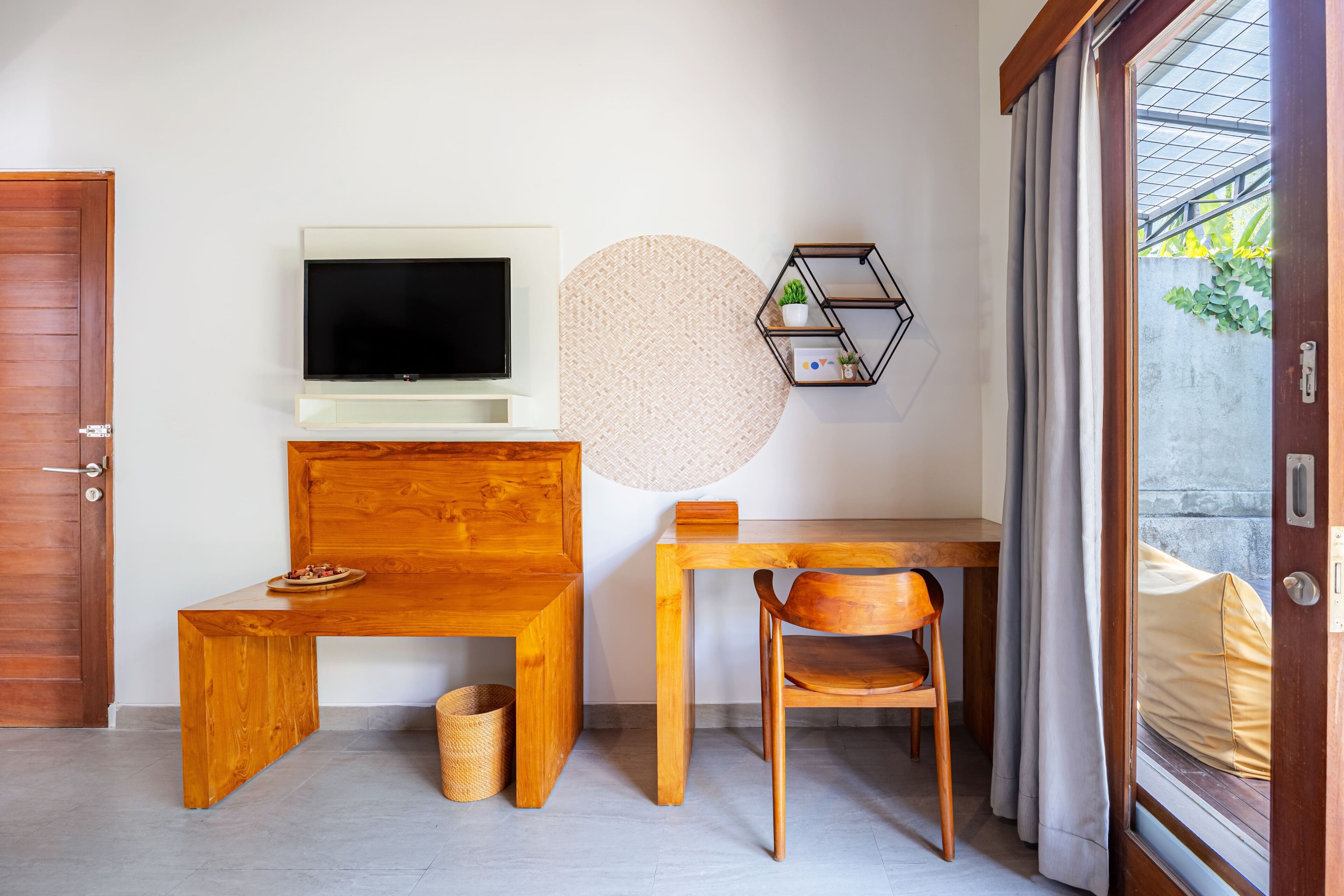 Une chambre moderne avec un bureau en bois, un téléviseur fixé au mur, un tapis rond en fibres naturelles et une étagère hexagonale ornée de plantes.