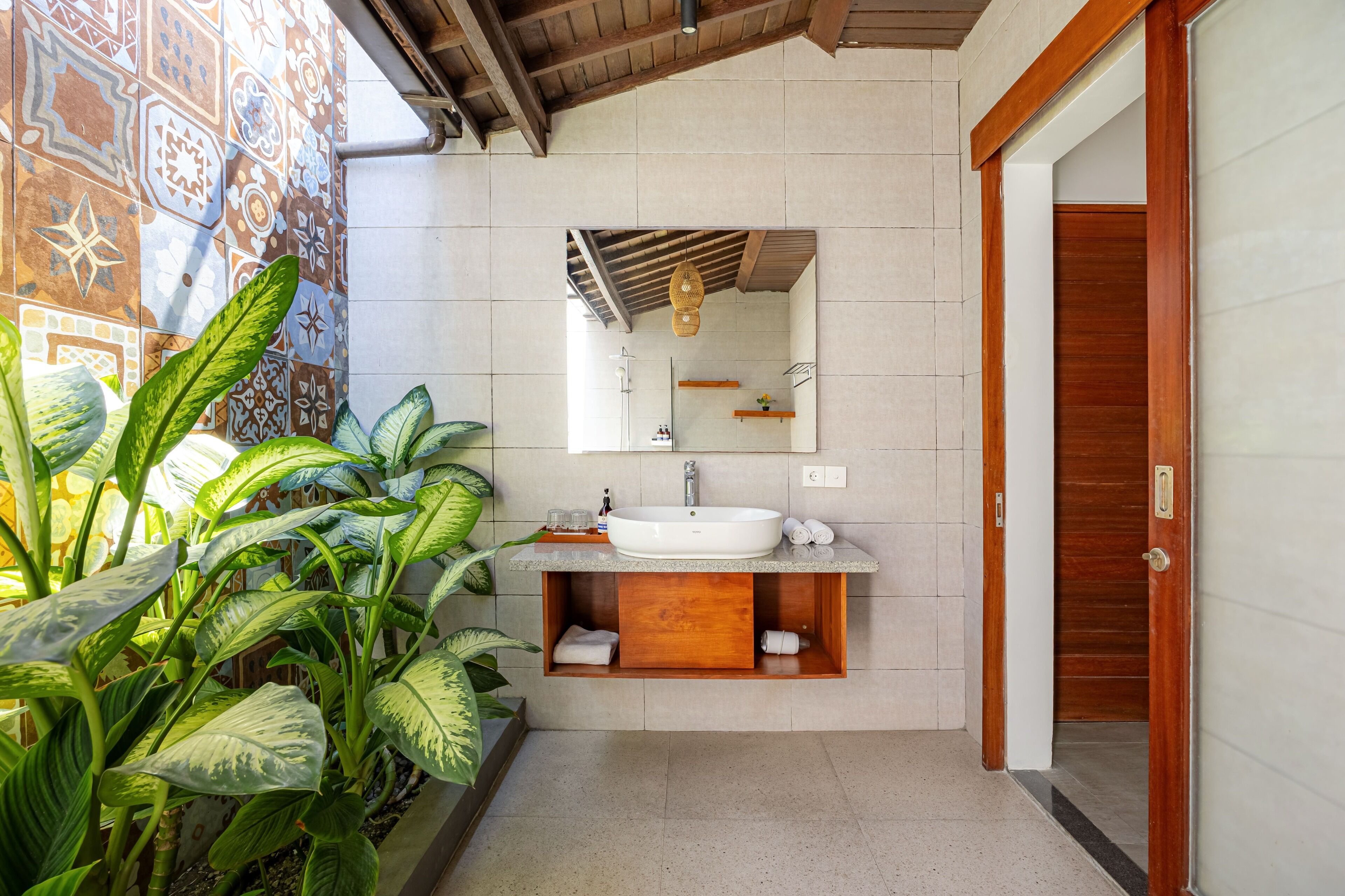 Un baño con un lavabo blanco, tocador de madera y un espejo encima. Hay plantas en el lado izquierdo y una puerta de madera a la derecha.