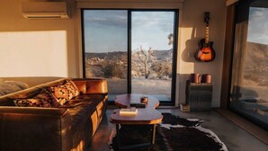 Un salón con un sofá de cuero, una mesa de centro redonda y un tapiz de piel de vaca. Una guitarra cuelga en la pared, y hay un ventanal con vistas a un paisaje desértico.
