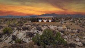 Un edificio moderno de un solo piso con techo plano, rodeado por un paisaje desértico rocoso con vegetación escasa.