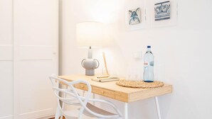 Een minimalistisch ingerichte kamer met een houten bureau, een witte stoel, een wandlamp en twee ingelijste kunstwerken.