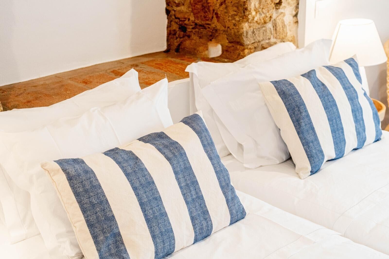 A bed with white bedding and striped pillows, a stone wall, and a lamp on a bedside table.