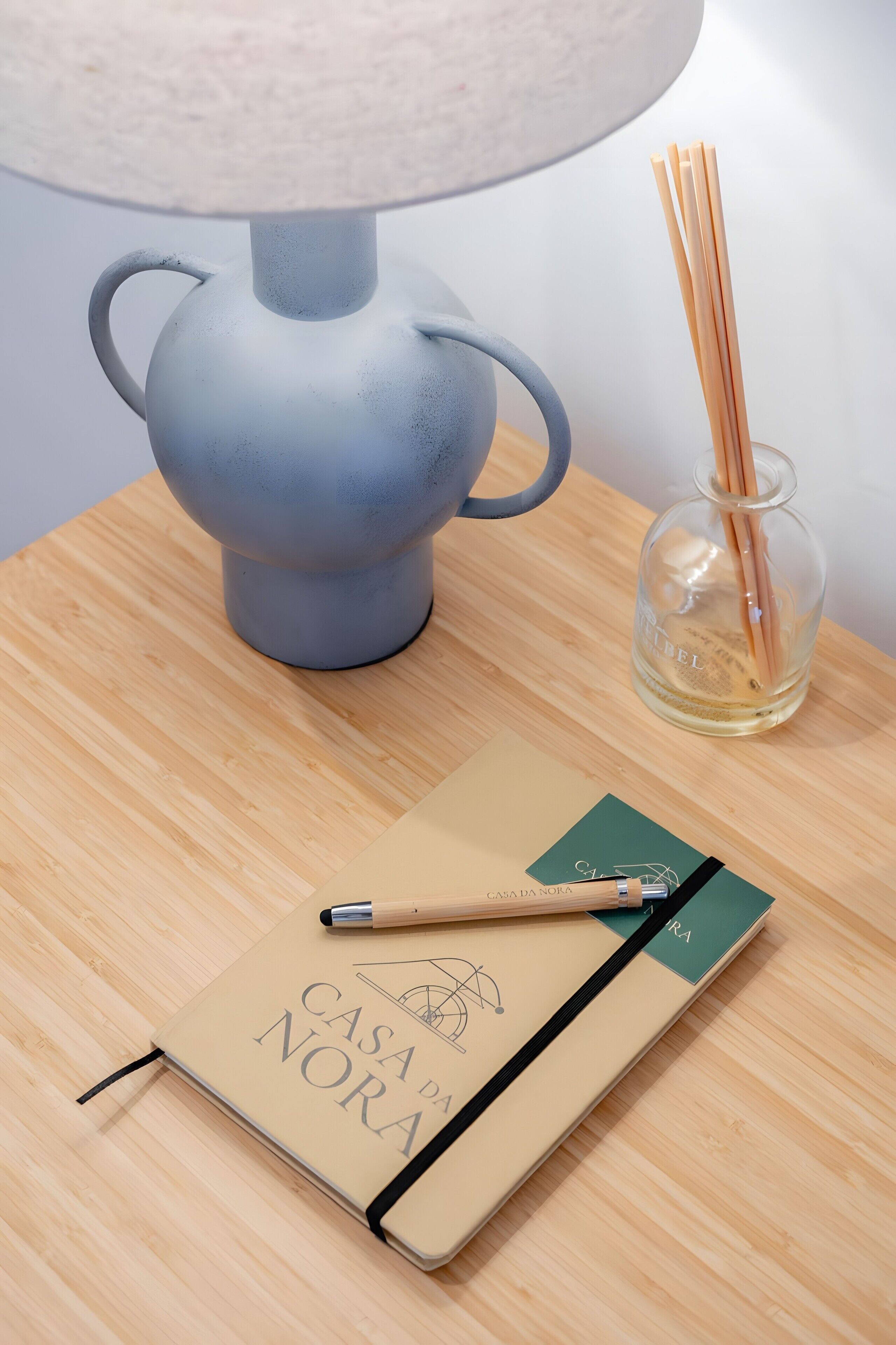 A notebook with 'CASA DA NORA' on the cover, a pen, a lamp, and a glass jar with reed sticks on a wooden table.