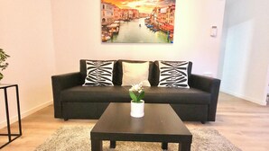 A living room with a black sofa, two patterned pillows, a white vase with flowers, and a painting of a canal scene on the wall. - Oshun Valdecanillas (Madrid)