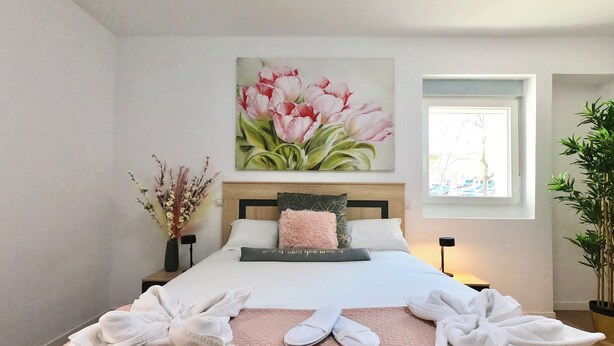 A neatly made bed with towels and slippers, a large floral painting above it, and a window with a view of the outside. - Oshun Valdecanillas (Madrid)