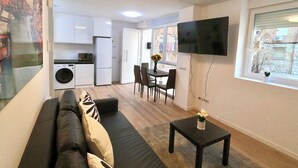A compact living space with a sofa, a small table, a TV, a dining area with chairs, a kitchen with appliances, and a window with a view. - Oshun Valdecanillas (Madrid)