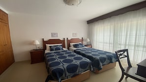 Quarto com duas camas, guarda-roupa de madeira, uma cadeira e uma escrivaninha.