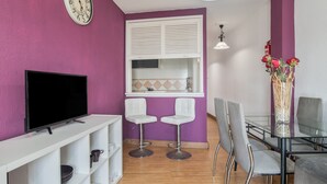 A modern living room with a flat-screen TV, white shelves, two white bar stools, a glass dining table with chairs, a white window with blinds, and a wall clock. - Apartment 50 Meters from the Beach with Terrace and Sea Views (Conil de la Frontera)