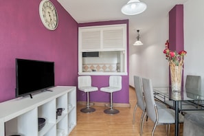 A modern living room with a flat-screen TV, white shelves, two white bar stools, a glass dining table with chairs, a white window with blinds, and a wall clock.