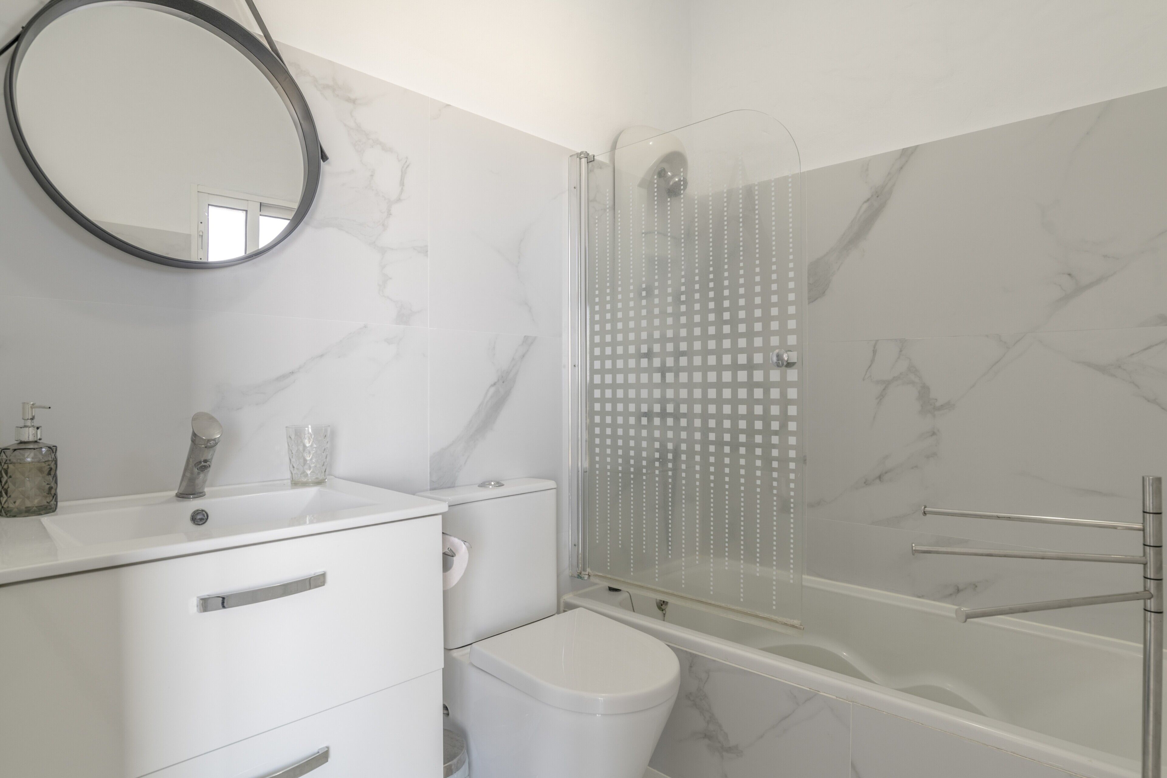 A bathroom with a marble wall, a sink with a mirror, a toilet, and a glass-enclosed shower.