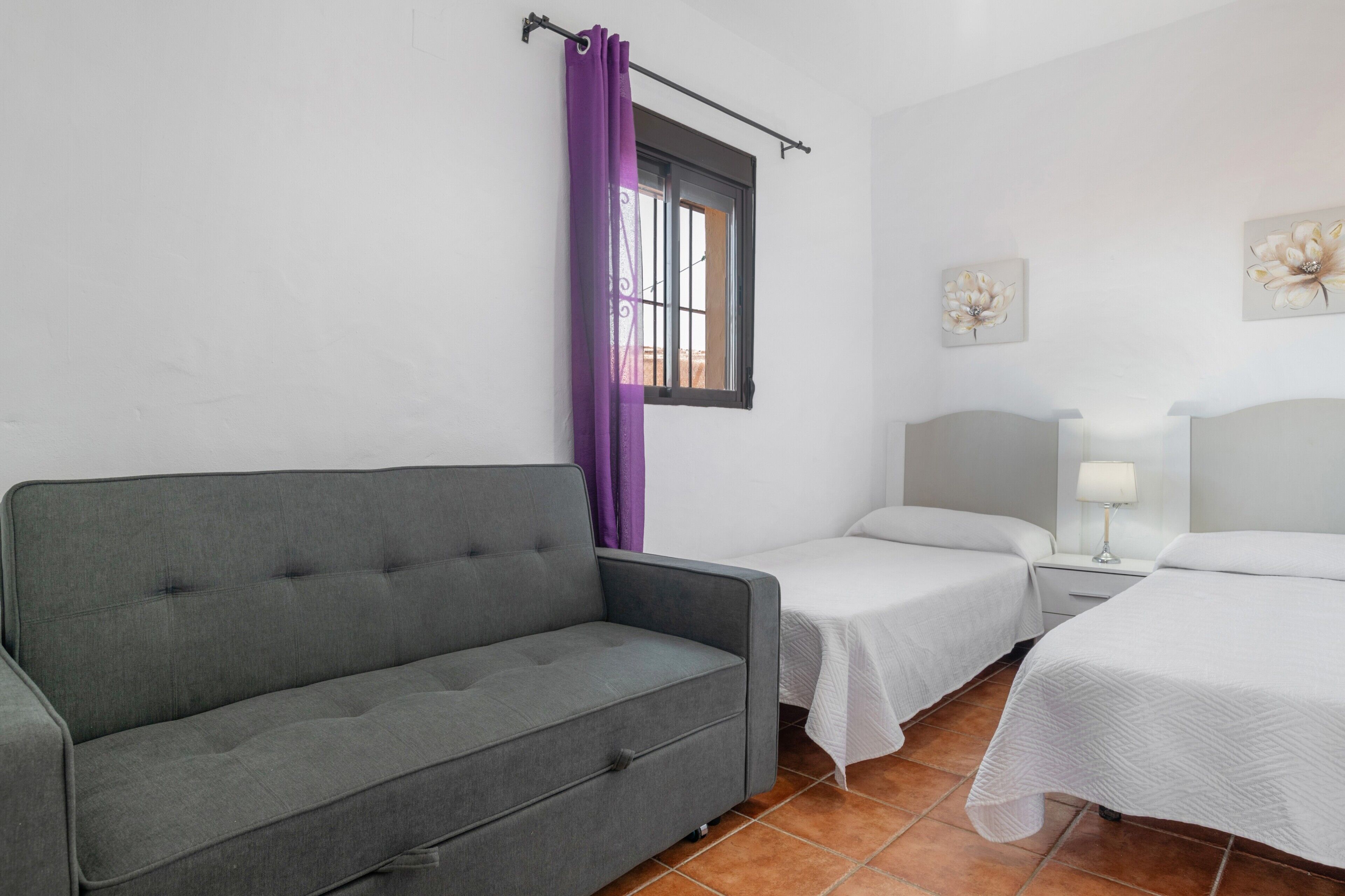 Una habitación con un sofá gris, dos camas individuales, un ventanal con cortinas moradas y dos cuadros de flores colgados en la pared.