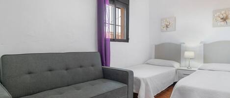 Una habitación con un sofá gris, dos camas individuales, un ventanal con cortinas moradas y dos cuadros de flores colgados en la pared.