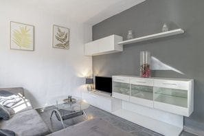 A modern living room with a sofa, a glass coffee table, a flat-screen TV, a white cabinet with glass doors, and two framed leaf artworks on the wall.
