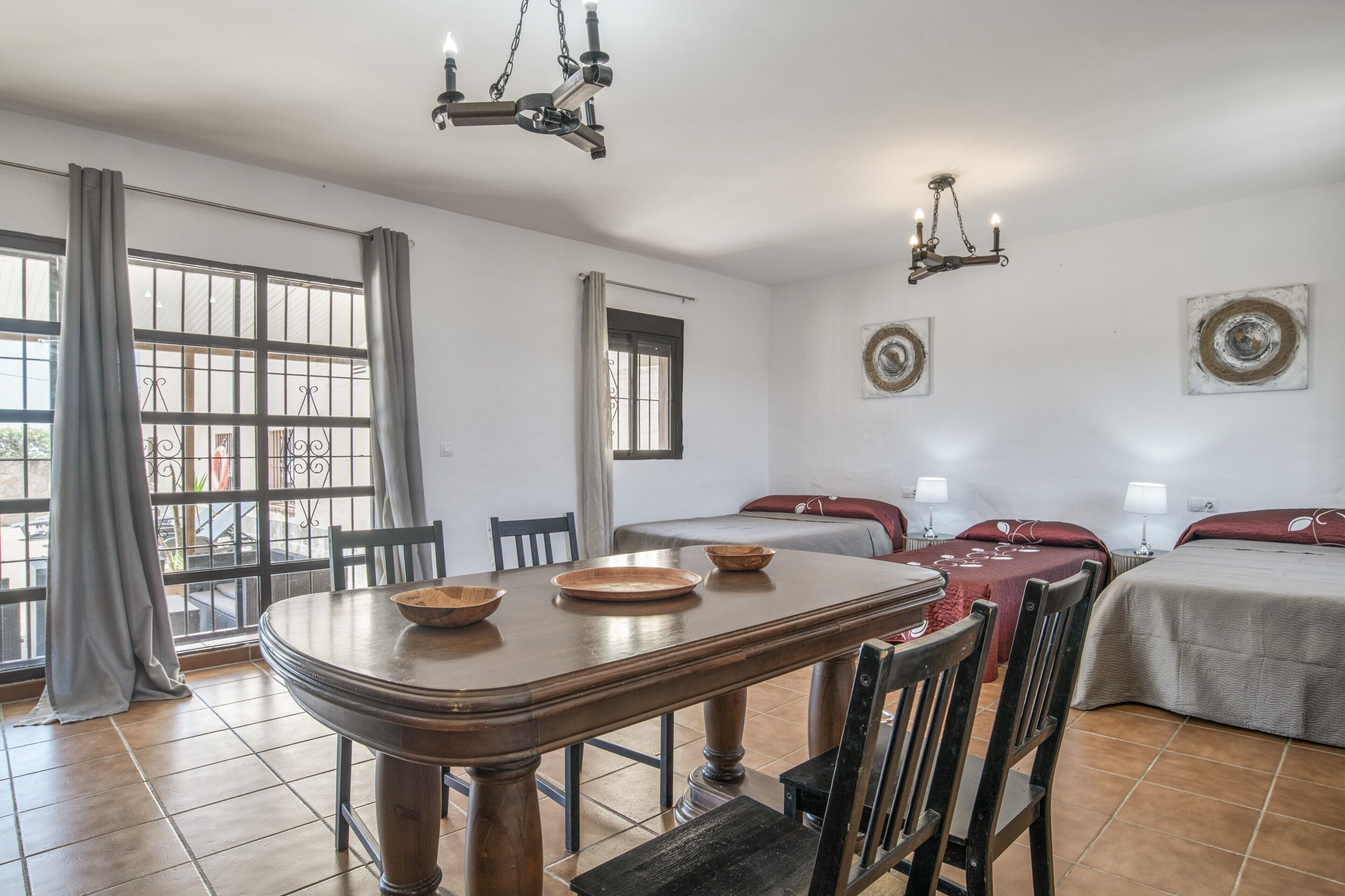 Una habitación con una mesa de comedor, sillas, una cama con cabecera roja, dos mesitas de noche con lámparas y un candelabro.