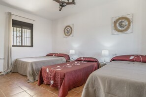 A hotel room with three beds, a window with bars, a ceiling fan, and two wall-mounted lamps.