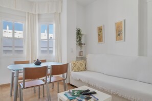 Una sala de estar con un sofá blanco, una mesa de comedor con sillas, una mesa de centro con libros y controles remotos, y una ventana con cortinas.