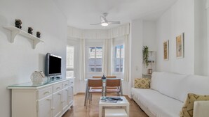 Una sala de estar con televisor, muebles blancos, ventilador de techo y comedor.