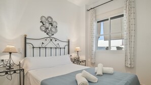Una cama bien hecha con una colcha azul, dos lámparas de noche, un adorno en la pared y una ventana con cortinas.