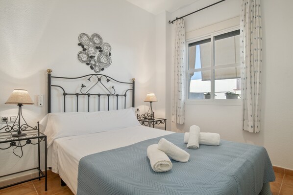 A neatly made bed with a blue bedspread, two bedside lamps, a decorative wall piece, and a window with curtains.