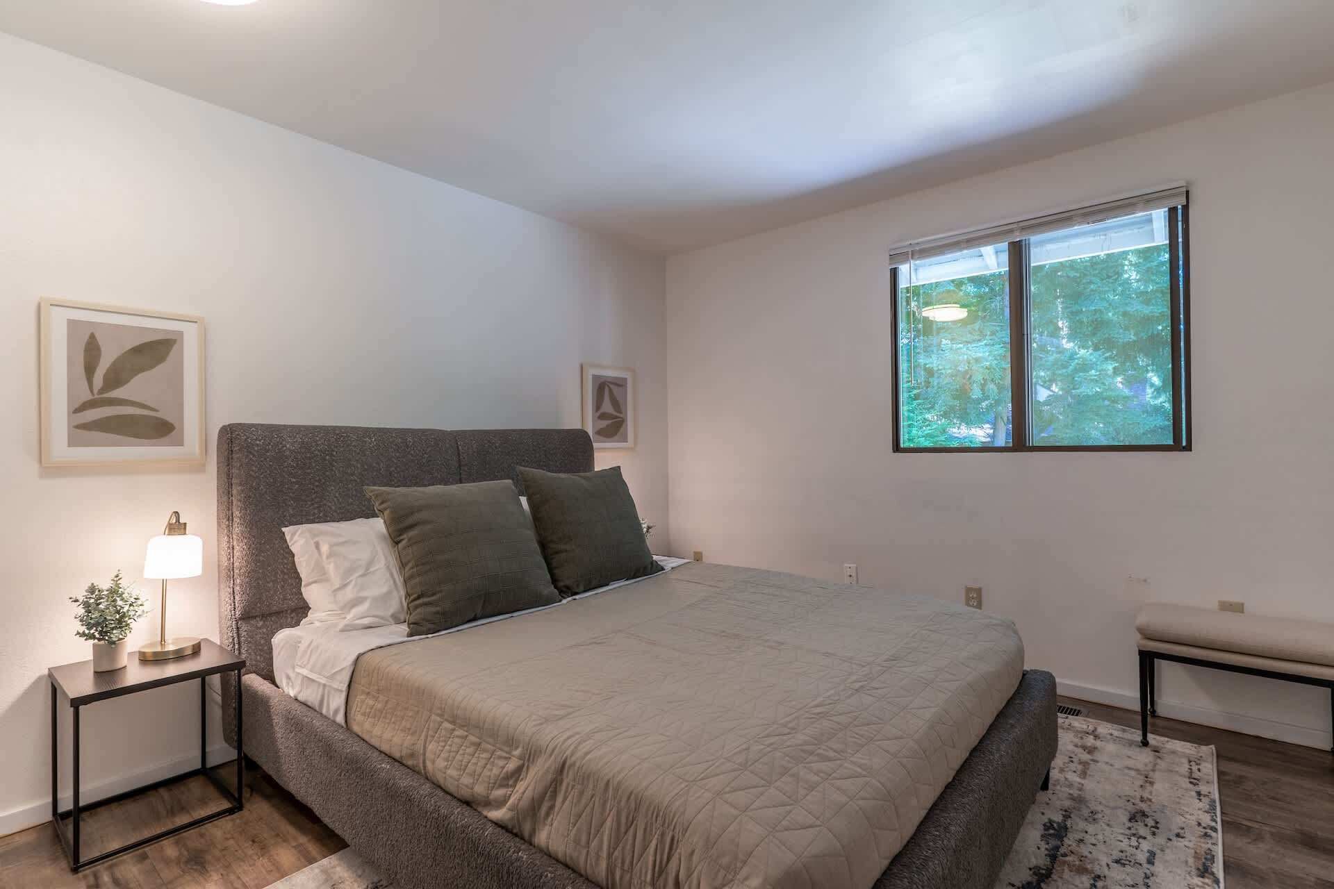 A bedroom with a bed, bedside tables, a lamp, a window with a view of trees, and a bench.