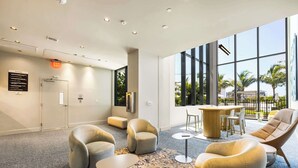 A modern lobby with seating, a reception desk, and large windows offering a view of palm trees and buildings.