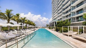 A swimming pool with lounge chairs and umbrellas, surrounded by palm trees and a modern building.