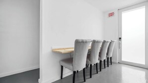 Ein schlicht eingerichteter Raum mit einem Holztisch und vier grau gepolsterten Stühlen. Ein Schild mit der Aufschrift EXIT ist an der Wand sichtbar.