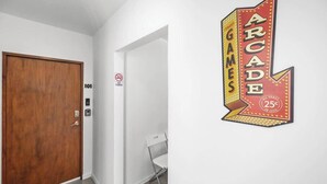 Ein Flur mit einer Holztür mit der Aufschrift '101', einer weißen Wand und einem Neonlichtschild, das 'Arcade Games' für 25 Cent bewirbt.