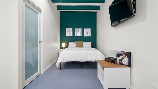 Ein schmaler Raum mit einem Bett, einem Schreibtisch und einem an der Wand befestigten Fernseher.