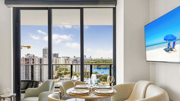 A modern hotel room with a large balcony, a dining table set for four, and a view of the cityscape and water.