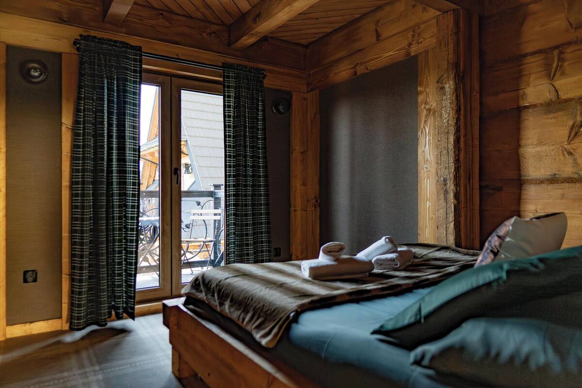 A wooden cabin bedroom with a bed, curtains, and a view of bicycles outside.