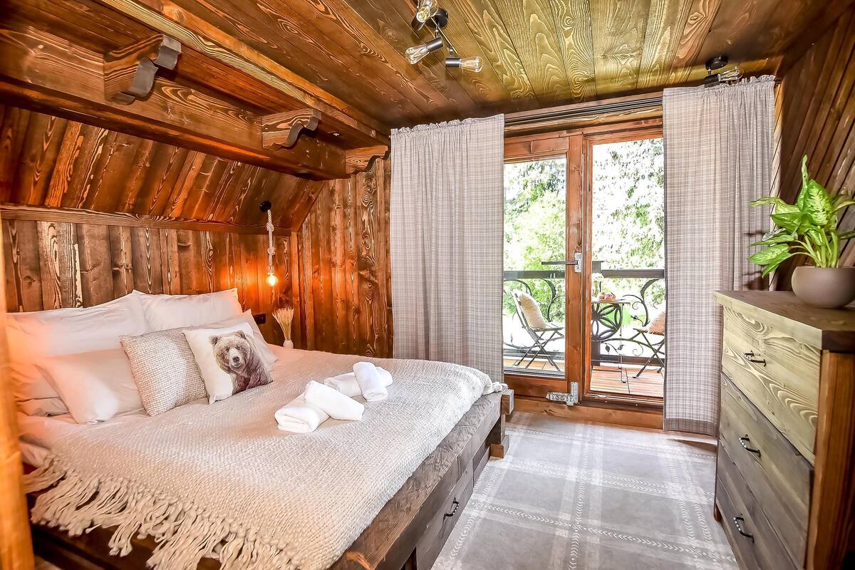 A wooden cabin bedroom with a bed, a dresser, and a balcony with outdoor furniture.