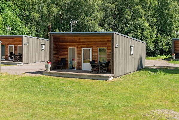 Une petite cabane à un seul étage, avec une façade en bois et un toit en métal, comprenant une terrasse avec une table et des chaises, ainsi qu’un petit jardin.