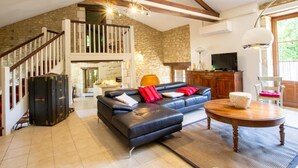 A spacious loft with exposed wooden beams, a stone wall, a black leather sofa, a dining table, and a staircase leading to an upper level. - Charming stone cottage with heated pool, pets welcome near Périgueux (Coulounieix-Chamiers)