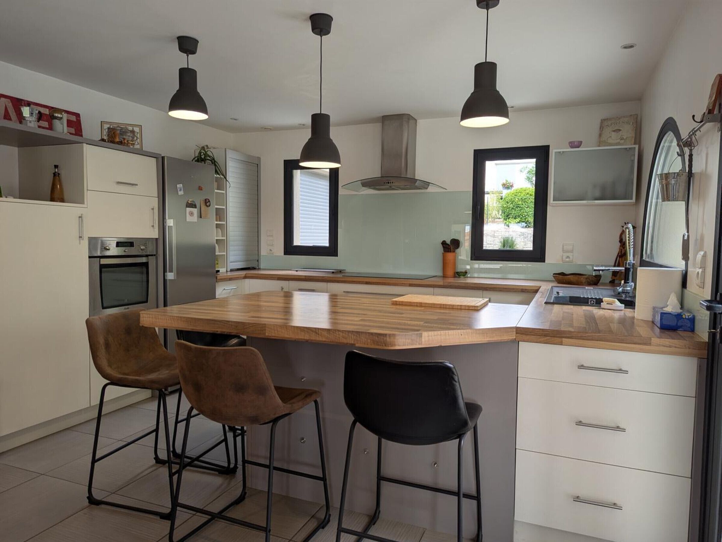 Une cuisine moderne dotée d’un îlot central, de tabourets de bar, de luminaires suspendus et offrant une vue sur l’extérieur grâce aux fenêtres.
