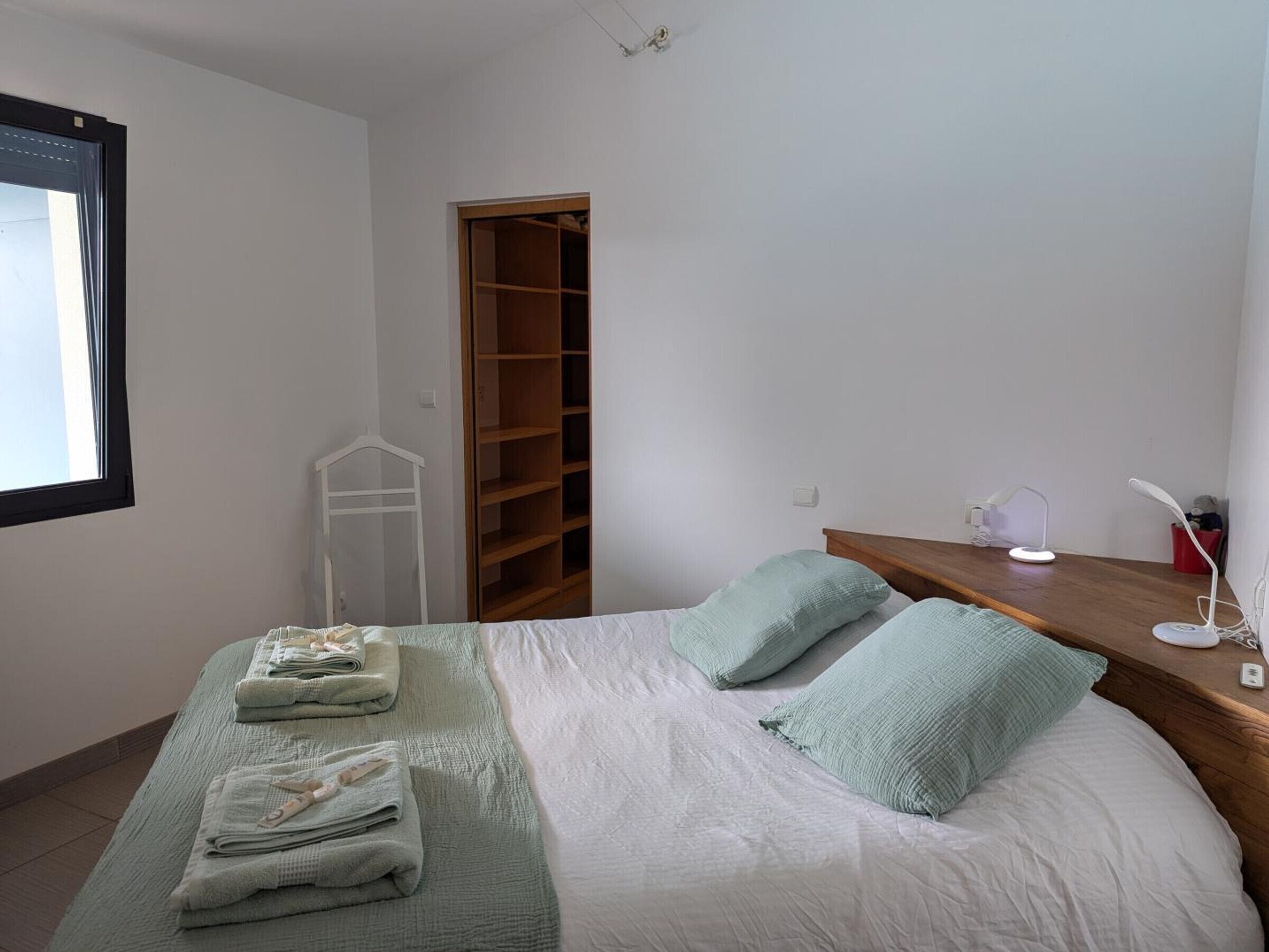 A bedroom with a bed, a wooden wardrobe, a bedside table with a lamp, and a window.