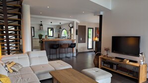 A modern living room with a sofa, ottoman, and coffee table. A kitchen with bar stools and a dining area with a TV and entertainment unit is visible in the background.