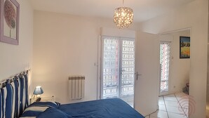 Un dormitorio con cama, mesita de noche, lámpara, lámpara de techo, radiador y una puerta que da a otra habitación.