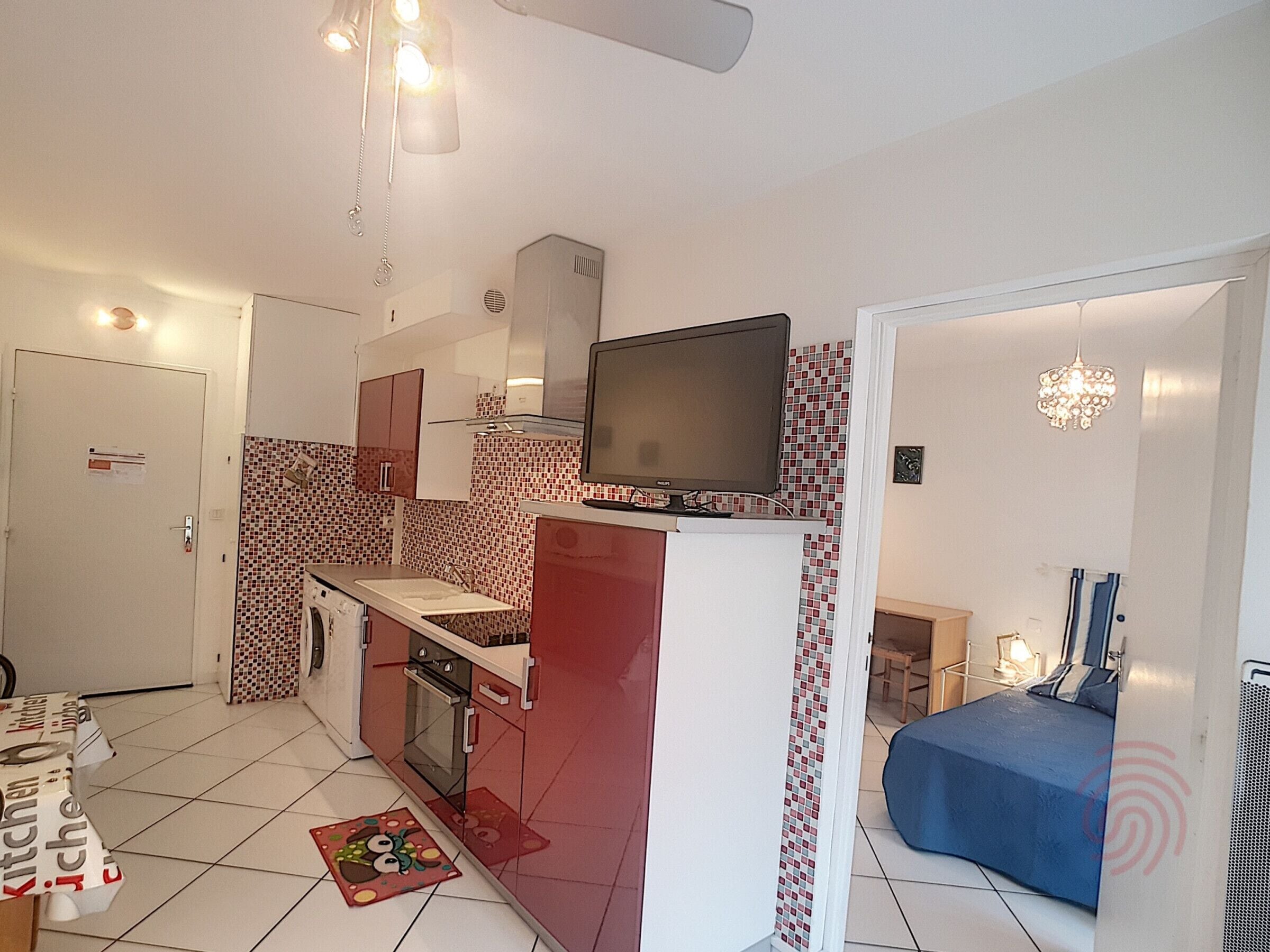 Uma cozinha compacta com televisão, micro-ondas e forno pequeno. Há uma mesa de jantar com uma cadeira e uma cama azul no quarto adjacente.