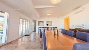 Een ruime eethoek met een lange houten tafel, blauwe gestoffeerde stoelen en een groot raam dat voor natuurlijk licht zorgt.