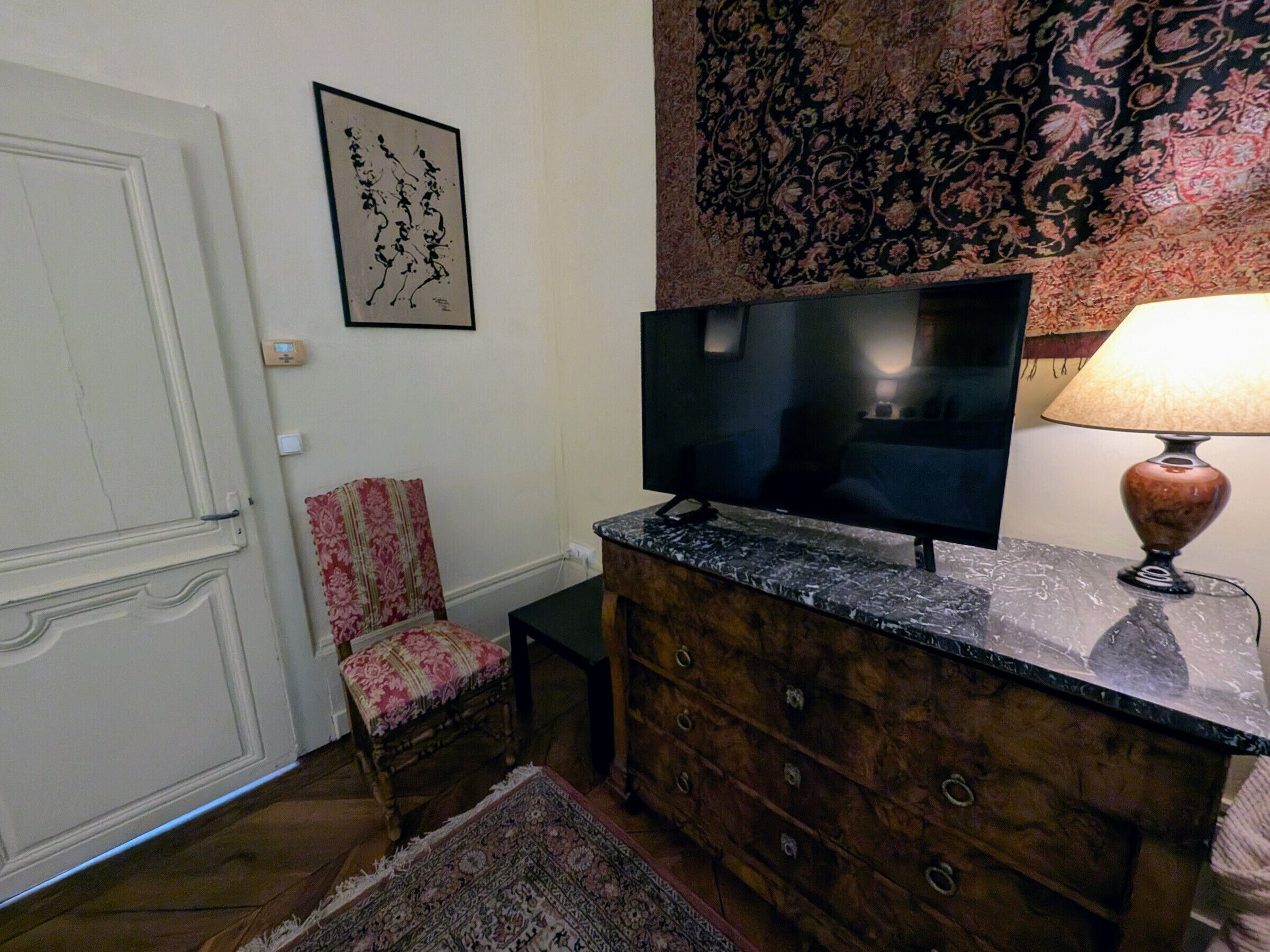 Una habitación con una televisión sobre un aparador de madera, una silla estampada, una alfombra y un adorno colgante en la pared.