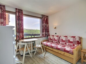 A cozy room with a wooden sofa, a small table, and a chair. A window with curtains offers a view of green hills.
