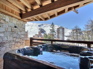 A hot tub with a stone wall and wooden beams, overlooking a snowy mountain and buildings.