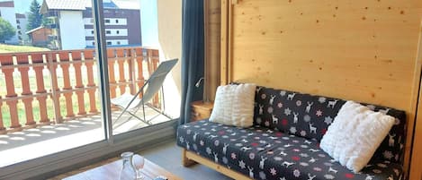 A cozy room with a wooden sofa, a small table with a glass, and a view of a balcony with a wooden fence.