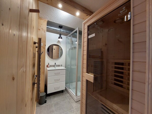 A modern bathroom with a wooden vanity, a round mirror, a glass shower enclosure, and a sauna door.